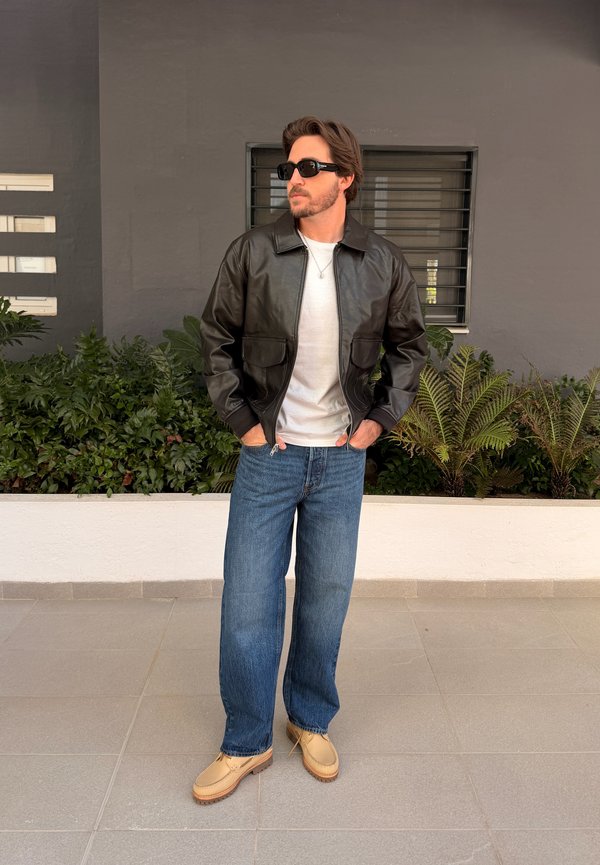 Hombre con chaqueta de cuero negra, camisa blanca, vaqueros azules y zapatos beige, de pie con las manos en los bolsillos cerca de plantas y una ventana.