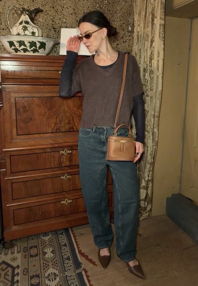 Mujer con gafas de sol, suéter marrón sobre camiseta negra, vaqueros holgados, zapatos marrones con punta y un pequeño bolso marrón cruzado, en el interior junto a una cómoda de madera.