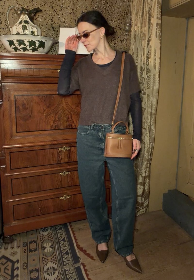 Kvinde iført solbriller, brun sweater lagdelt over sort top, løse jeans, brune spidse sko og en lille brun crossbody taske indendørs ved et trækøjleskab.