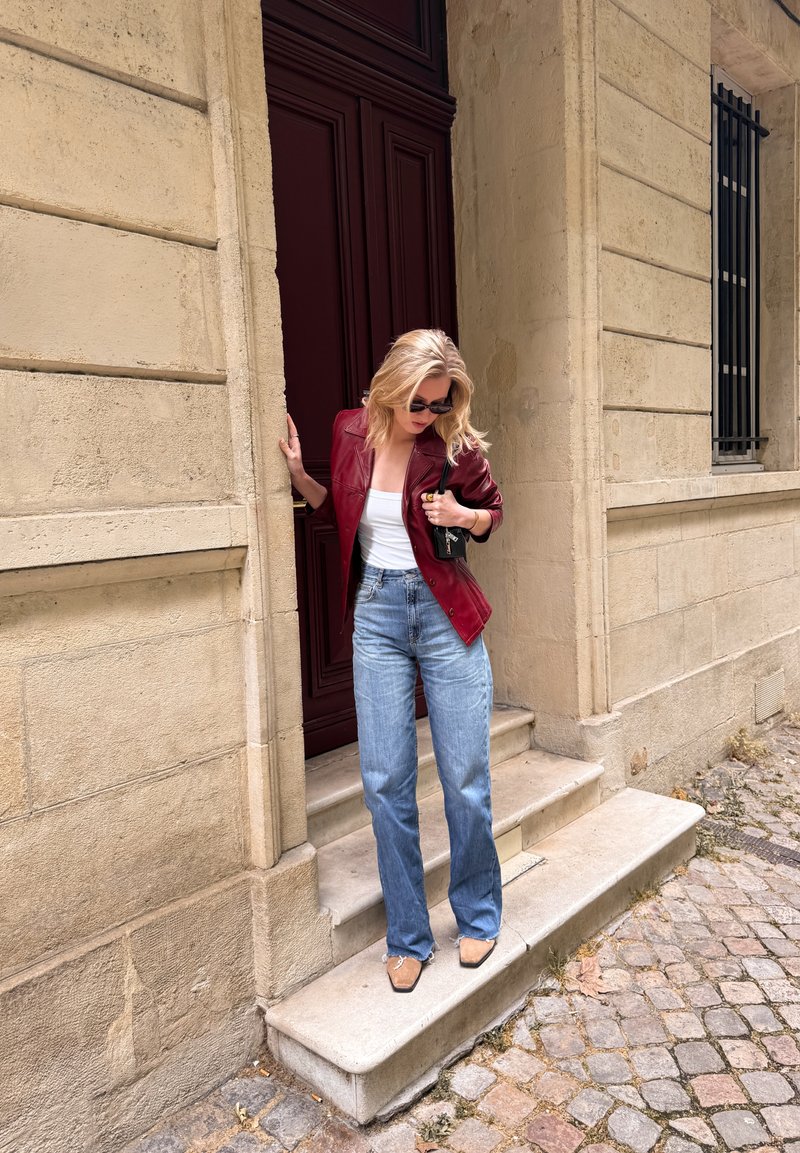 Mujer con gafas de sol bajando escaleras de piedra, lleva una chaqueta de cuero roja, camiseta blanca, vaqueros azules y zapatos beige en una calle empedrada.