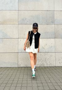 Svart ermeløs vest over en hvit topp, sammen med et hvitt skjørt. Grønn sneakers med rosa detaljer, solbriller og en svart caps. Betongbakgrunn.