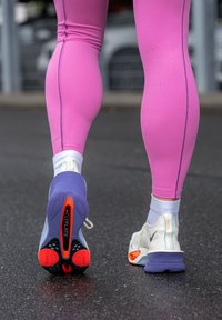 Scarpe da corsa leggere con tomaia in mesh bianco e dettagli viola, arancioni e neri, abbinate a leggings rosa aderenti.