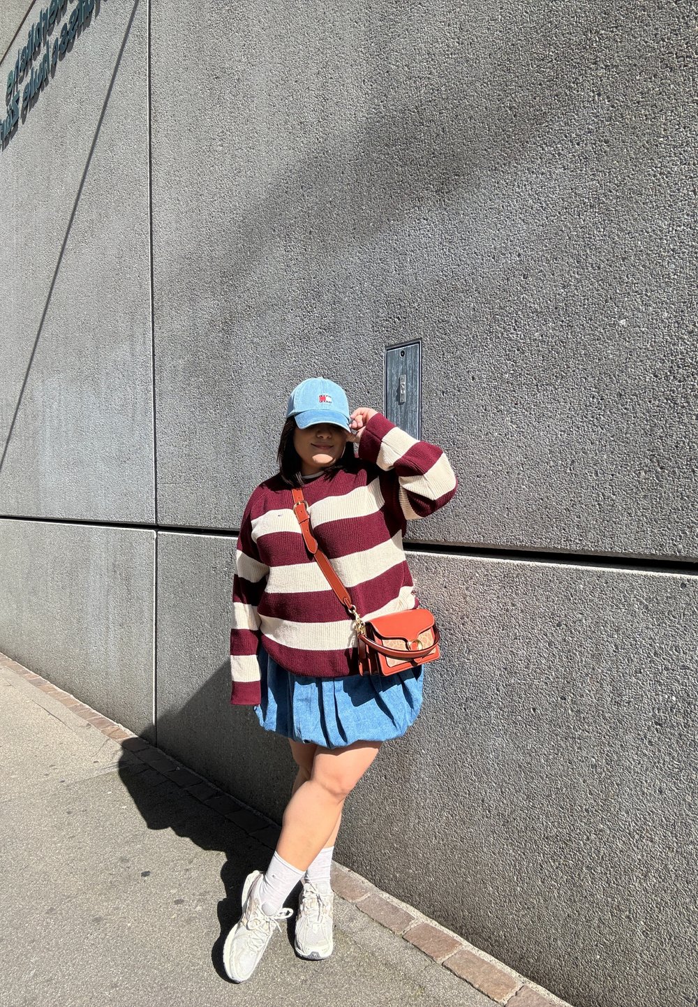 Maglione a righe bordeaux e crema, gonna di jeans, cappello azzurro chiaro, piccola borsa arancione a tracolla, calze bianche e sneakers, davanti a un muro di cemento.