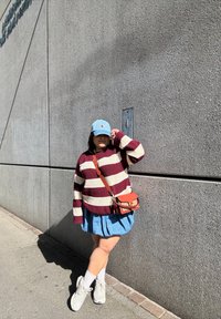 Maglione a righe bordeaux e crema, gonna di jeans, cappello azzurro chiaro, piccola borsa arancione a tracolla, calze bianche e sneakers, davanti a un muro di cemento.