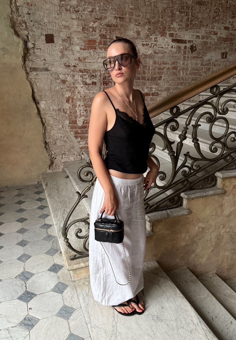 Femme portant un haut noir sans manches, une jupe blanche, de grandes lunettes de soleil et des sandales noires, tenant un sac noir, debout près d'un escalier en marbre avec une rampe en fer.