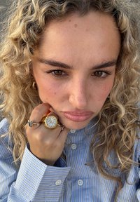 Zbliżenie na kobietę z kręconymi blond włosami, ubraną w koszulę w paski, noszącą złoty zegarek i pierścionek, trzymającą rękę blisko twarzy.