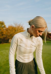 Hellbeiges Langarmshirt mit Reißverschluss und taillierter Passform, mit strukturierter Stoffstruktur und dezentem Branding, kombiniert mit dunklen Hosen mit elastischem Bund.