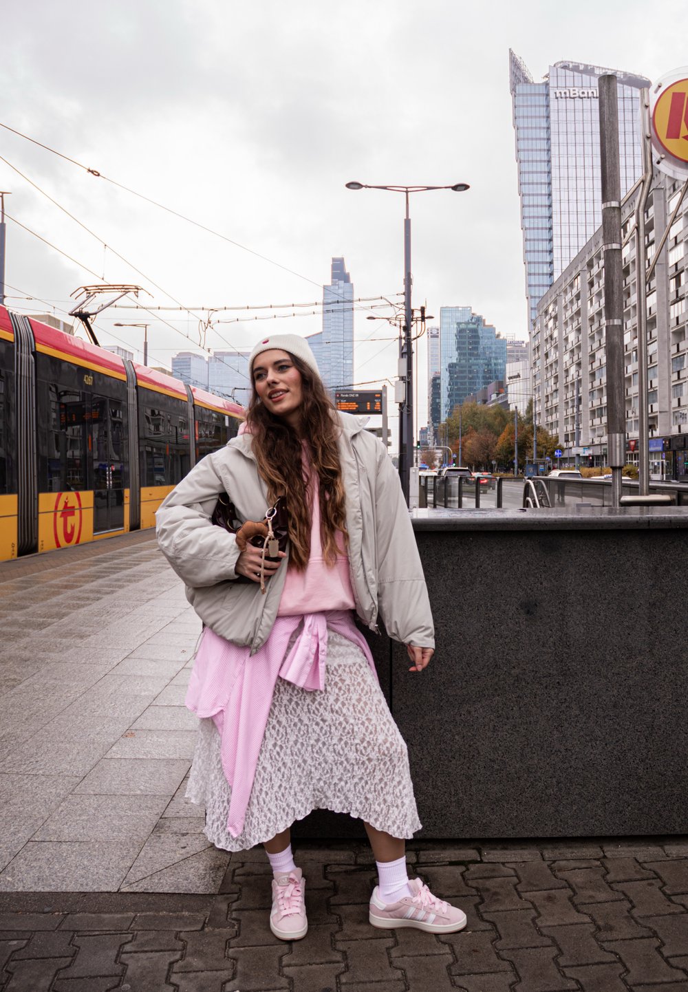 Lichtgrijze pufferjack over een roze sweatshirt, gecombineerd met een witte kanten rok en roze sneakers. De achtergrond toont een stedelijk landschap en een tram.