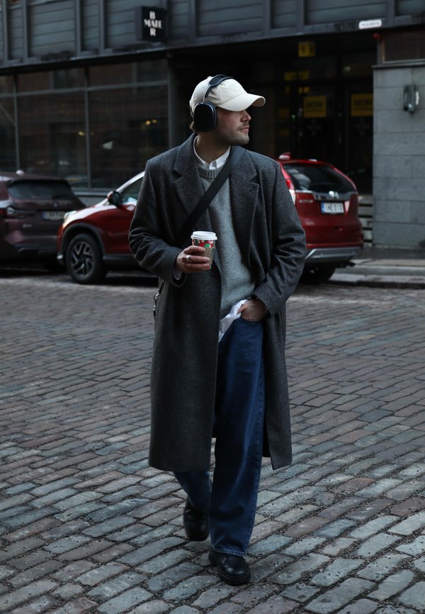 Cappotto di lana grigio, maglione grigio, camicia bianca, pantaloni in denim blu, scarpe nere. Indossa cuffie e tiene una tazza di caffè. Strada acciottolata.