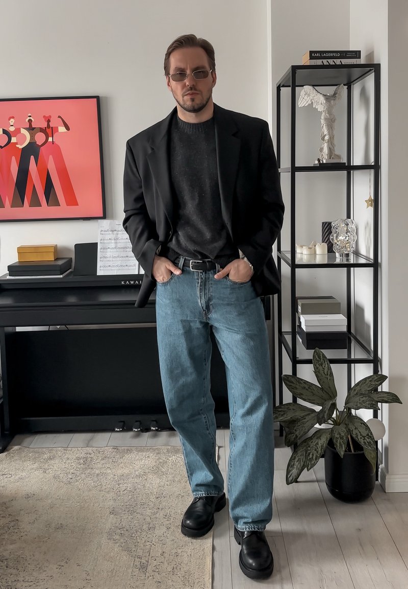 Blazer negro sobre un suéter oscuro, jeans azul claro y zapatos negros. De fondo hay una obra de arte rosa y una estantería negra con decoración.