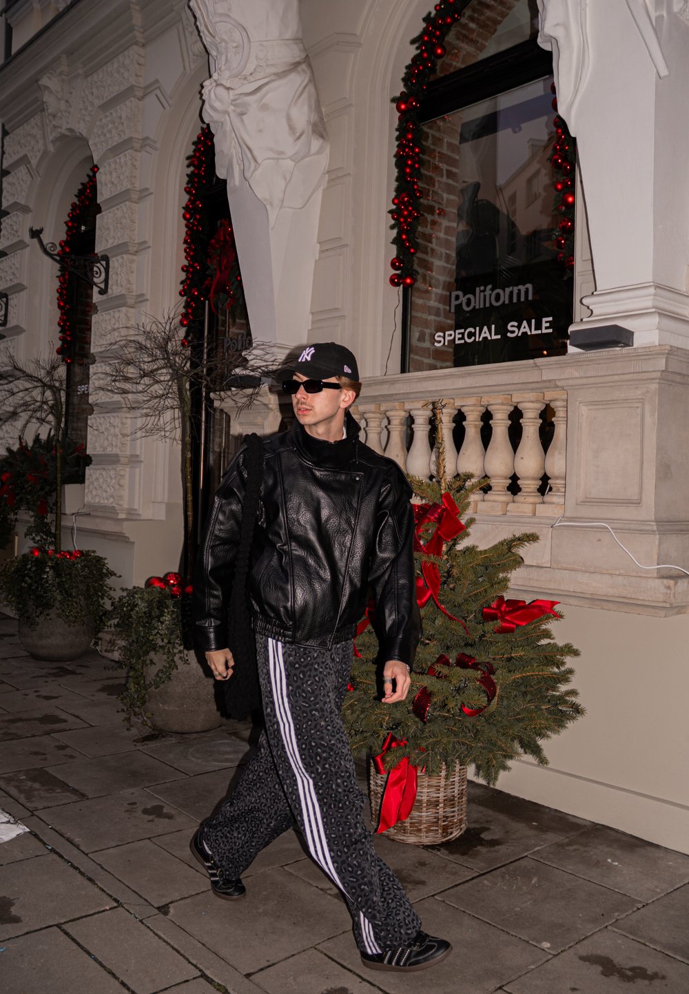 Veste en cuir noire sur un haut noir, pantalon large à motifs avec des rayures blanches, casquette foncée et lunettes de soleil, marchant devant une vitrine décorée.