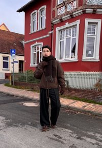 Ruda, izoliuota striukė ant juodo aprangos komplekto, puošta tamsia skarele su kutais. Fone matyti raudonas pastatas su baltomis apdailos detalėmis.