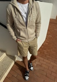 Beige zip-up hoodie with fleece lining, white logo, paired with light khaki shorts and black/white sneakers on a brick surface.
