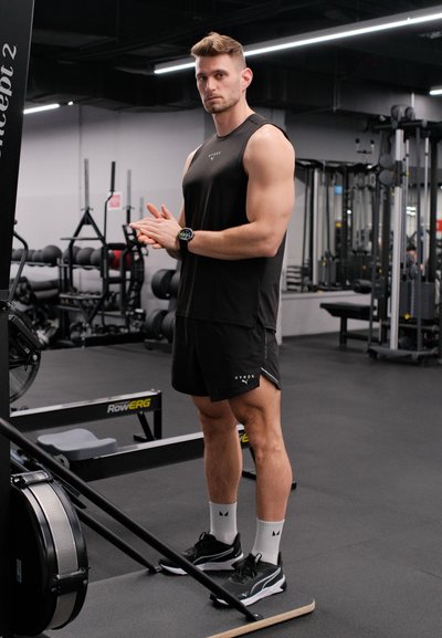 Hombre musculoso con ropa negra de entrenamiento de pie junto a una máquina de remo en un gimnasio, preparándose para hacer ejercicio.