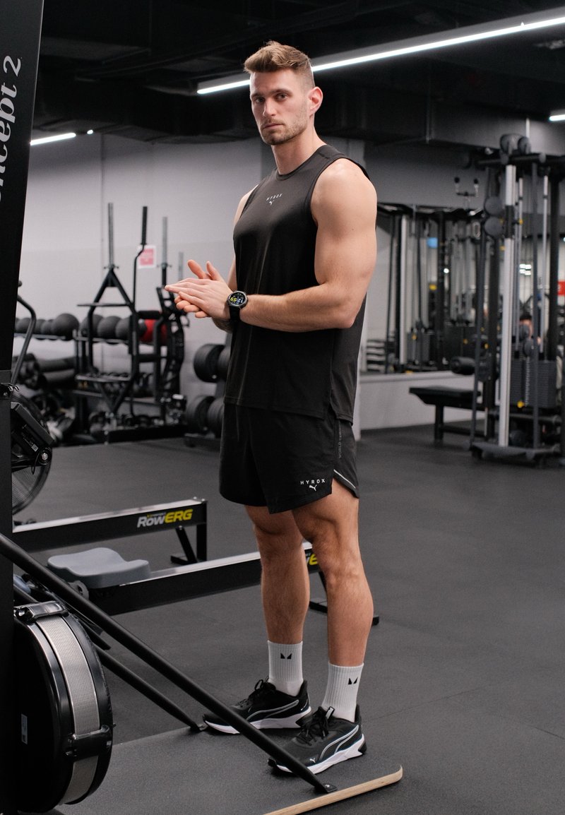 Homme musclé en vêtements de sport noirs debout à côté d'un rameur dans une salle de sport, prêt à faire de l'exercice.