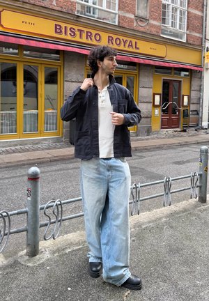 Joven ajustando una chaqueta negra mientras está de pie en la calle frente al Bistro Royal amarillo con toldo rojo y acera de adoquines.