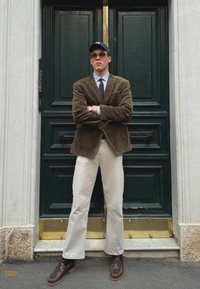 Blazer di velluto a coste marrone, camicia azzurra chiara, cravatta blu navy, pantaloni crema, scarpe marroni. In piedi davanti a una porta verde con dettagli dorati.