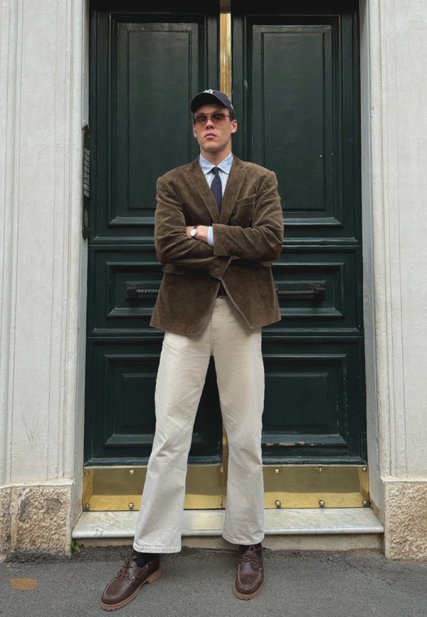 Blazer di velluto a coste marrone, camicia azzurra chiara, cravatta blu navy, pantaloni crema, scarpe marroni. In piedi davanti a una porta verde con dettagli dorati.