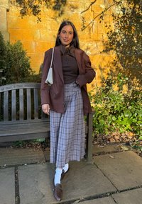 Blazer marron cintré porté sur un haut marron foncé, assorti à un pantalon large à carreaux gris et des mocassins marron. Accessoirisé d'un petit sac.