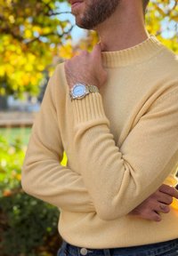 Άνδρας με γένια σε μπεζ πουλόβερ να αγγίζει τον λαιμό του, φοράει ασημένιο ρολόι καρπού, στέκεται σε εξωτερικό χώρο με φόντο τα φθινοπωρινά φύλλα.