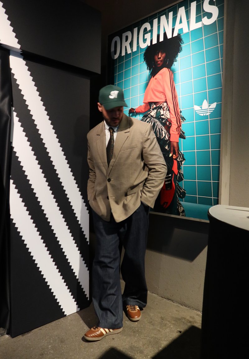 Homme en blazer beige, casquette verte et chaussures Adidas marron se tient près d'un panneau à rayures noir et blanc et d'une affiche colorée « ORIGINALS ».