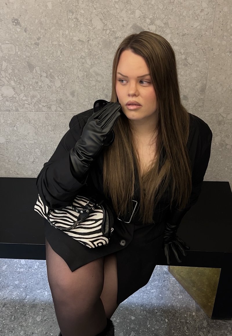 Kobieta o długich brązowych włosach, ubrana w czarny płaszcz i czarne rękawiczki, trzymająca torebkę w paski zebry, siedzi na czarnej ławce przy kamiennej ścianie.