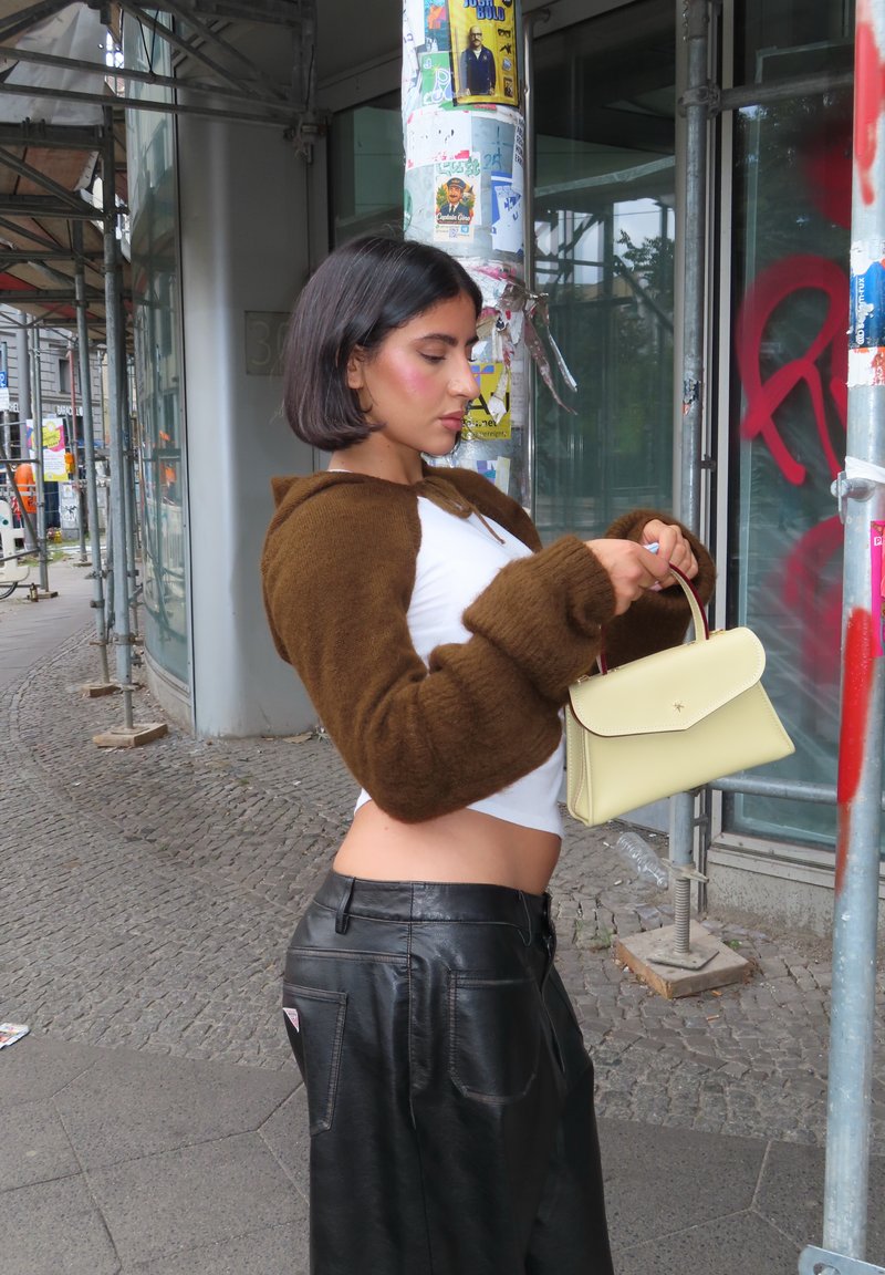 Brauner, gekürzter Pullover mit Puffärmeln; weißes eng anliegendes Oberteil; schwarze Lederhose; gelbe Handtasche mit strukturiertem Design; urbaner Hintergrund.