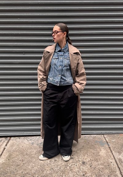 Abrigo largo beige sobre una chaqueta de mezclilla, pantalones anchos negros y zapatillas en blanco y negro. Lleva gafas y el pelo recogido, de pie contra una pared gris.