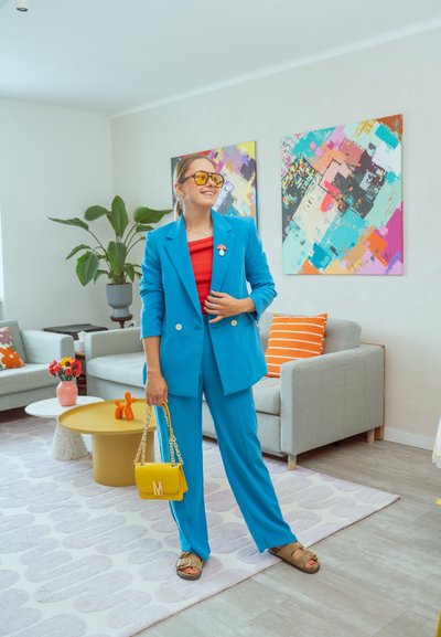 Conjunto azul brillante a medida con chaqueta cruzada y pantalones a juego, combinado con una blusa roja. Los accesorios incluyen un bolso amarillo y gafas de sol.