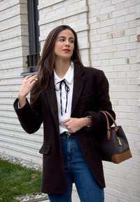 Femme aux longs cheveux bruns ajuste sa coiffure tout en portant un manteau sombre, un chemisier blanc avec une cravate noire, un jean bleu, et portant un sac à main marron en extérieur.