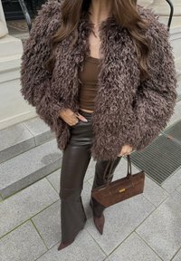 Palton maro din blană artificială pufoasă; maieu maro conținut; pantaloni evazați maro din piele; geantă mică maro cu accesorii aurii.