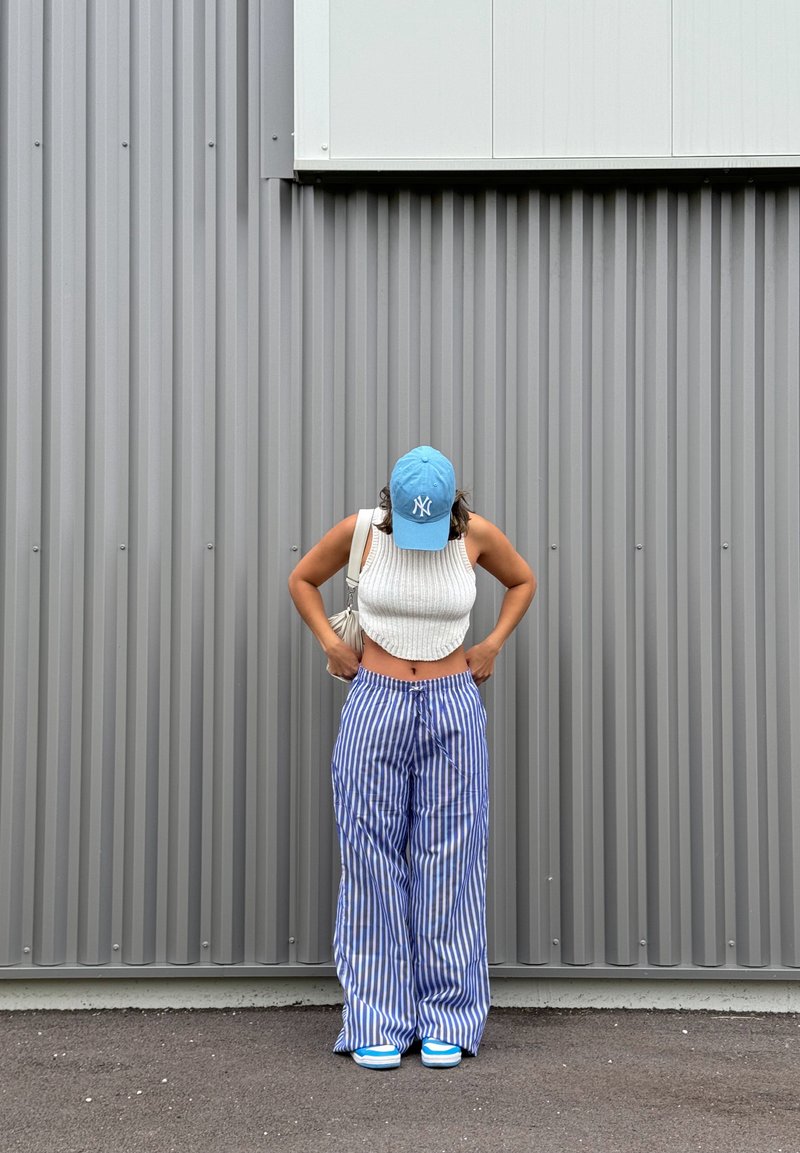 Λευκό ριγωτό τοπ με κοντά μανίκια, μπλε καπέλο μπέιζμπολ με λογότυπο, μπλε και λευκό ριγέ παντελόνι με φαρδιά πόδια και μπλε αθλητικά παπούτσια. Φόντο με γκρίζα κυματοειδή τοιχοποιία.