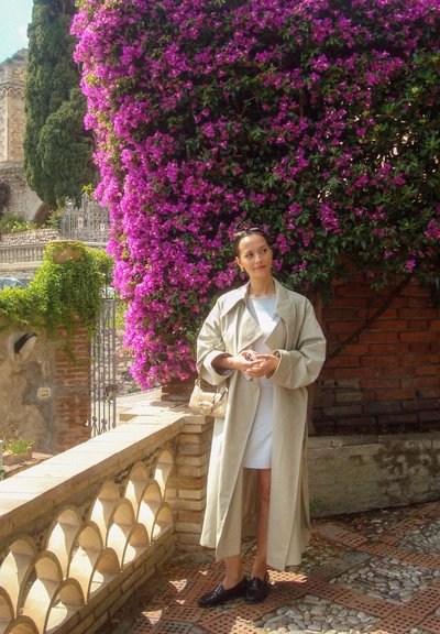 Gabardina beige, vestido blanco, mocasines negros. Rodeada de vibrantes bugambilias moradas y un muro de piedra con arcos decorativos.