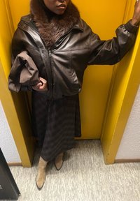 Hnědá kožená bomber bunda s kožešinovým límcem, nošená přes vzorované tmavé šaty, doplněná béžovými kozačkami nad kolena, na žlutém pozadí.