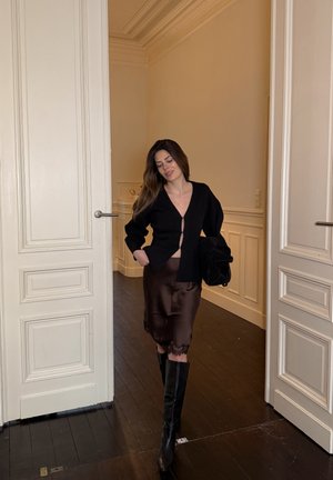 Mujer caminando en el interior entre puertas blancas dobles, vestida con un cárdigan negro, falda de seda oscura y botas negras hasta la rodilla, sosteniendo una chaqueta negra.