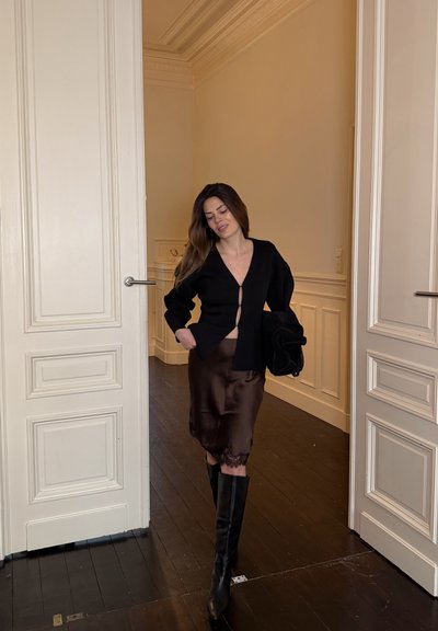 Mujer caminando en el interior entre puertas blancas dobles, vestida con un cárdigan negro, falda de seda oscura y botas negras hasta la rodilla, sosteniendo una chaqueta negra.