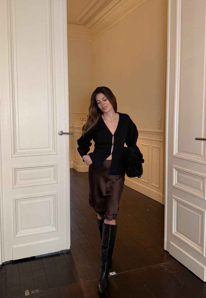 Donna che cammina all'interno tra porte doppie bianche, indossa un cardigan nero, una gonna di seta scura e stivali neri al ginocchio, tenendo una giacca nera.