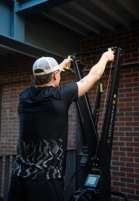 Mees, kes kannab valget nokamütsi ja musta särk ning kasutab siseruumis tellisseina taustal Concept2 SkiErg masinat.