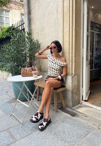 Top branca de ombro caído com bolinhas pretas, saia mini castanha, sapatos escorregões com padrão de vaca e uma mala castanha em uma mesa branca.