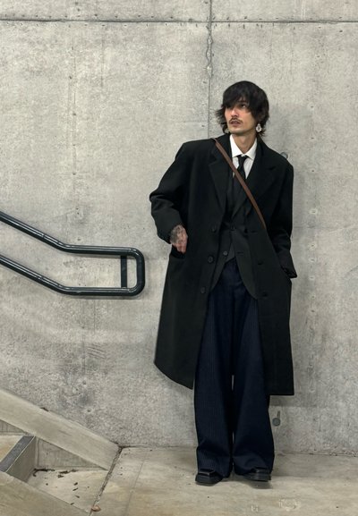 Abrigo negro sobre un traje a medida, pantalones anchos a rayas y un bolso marrón de hombro. Pared de concreto al fondo.