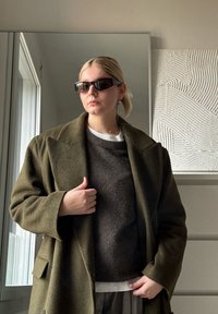 Olivengrüner Wollmantel mit breiten Revers, über einem dunkelbraunen Strickpullover, grauen Karo-Hosen und schwarzen rechteckigen Sonnenbrillen kombiniert.