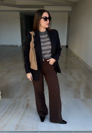Mujer con blazer negro, suéter gris a rayas, pantalones anchos marrón oscuro, zapatos negros, gafas de sol y llevando un bolso de hombro tejido color marrón claro en interiores.