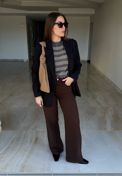 Mujer con blazer negro, suéter gris a rayas, pantalones anchos marrón oscuro, zapatos negros, gafas de sol y llevando un bolso de hombro tejido color marrón claro en interiores.