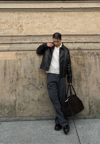 Μαύρο δερμάτινο μπουφάν πάνω από ένα κρεμ πλεκτό πουλόβερ, συνδυασμένο με γκρι παντελόνι με ρίγες. Κρατάει μια σκούρα τσάντα, στέκεται δίπλα σε έναν υφασμάτινο τοίχο.