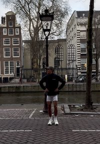 Personne athlétique en veste noire et short blanc se tient près d'un lampadaire. En arrière-plan, des bâtiments historiques et un canal.