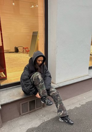 Mujer con una chaqueta negra con capucha y pantalones de camuflaje sentada en un bordillo, atándose el zapato negro de Nike en una calle de la ciudad.