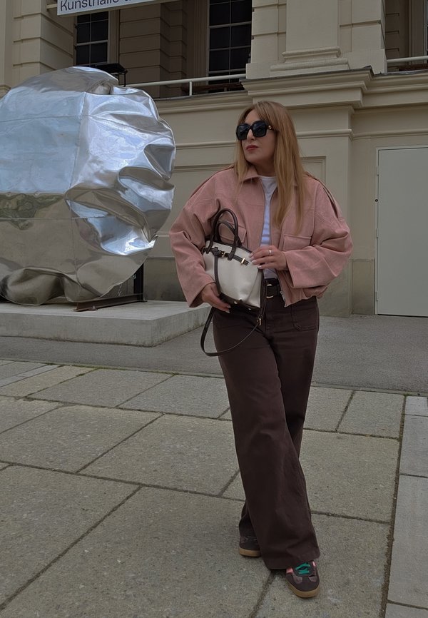 Žena s sunčanim naočalama u ružičastoj jakni i smeđim hlačama stoji blizu velike sjajne apstraktne metalne skulpture ispred zgrade.
