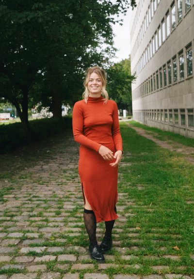 Vestido largo, ajustado y de punto acanalado en color naranja quemado con aberturas laterales. Combinado con medias negras transparentes y zapatos negros. El fondo presenta vegetación y un edificio.