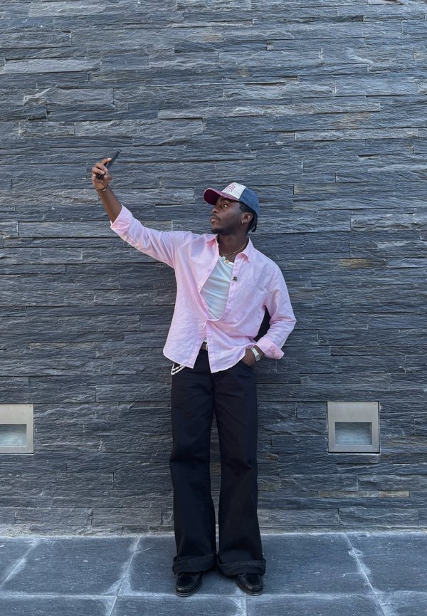 Person wearing a light pink button-up shirt, white t-shirt, black wide-leg trousers, and a grey cap with red accents, posing against a stone wall.