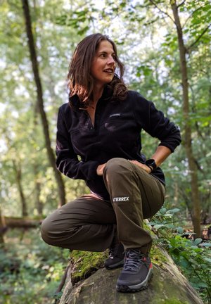 Sudadera de forro polar negra, pantalones cargo verdes y zapatos de senderismo grises con acentos rojos. Entorno natural, modelo agachado sobre un tronco.